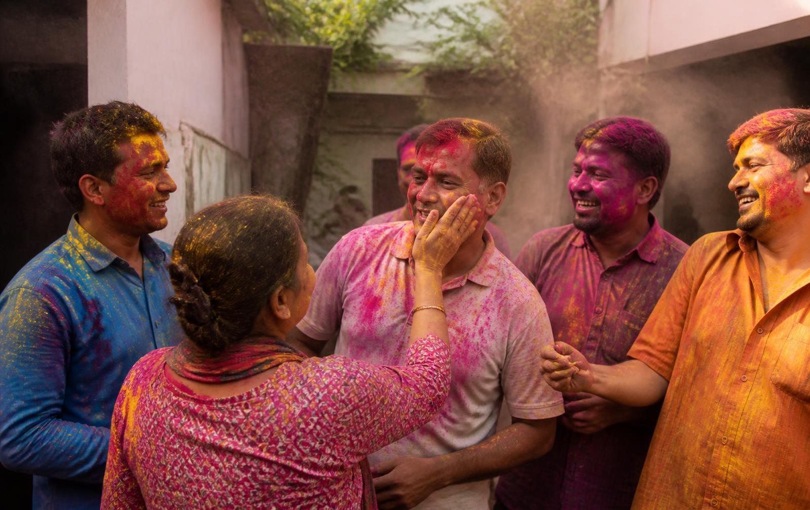 A small group of adults laughing together during Holi in a neighbourhood courtyard, dusted with coloured powder